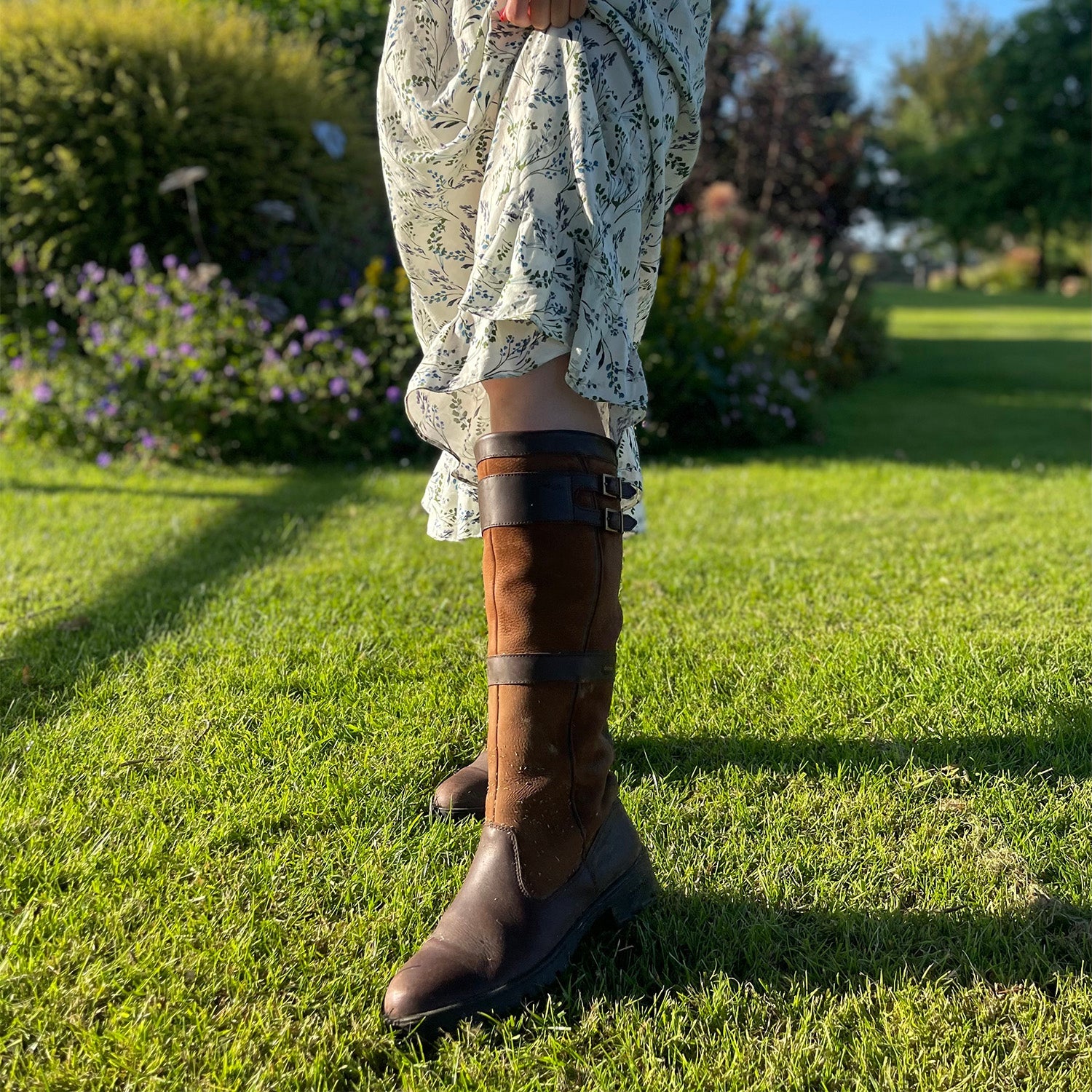 Dubarry Longford Leather Waterproof and Breathable Country Boot being worn with a dress on a well-maintained lawn with some shrubs in the background.//© Bonnie Baker