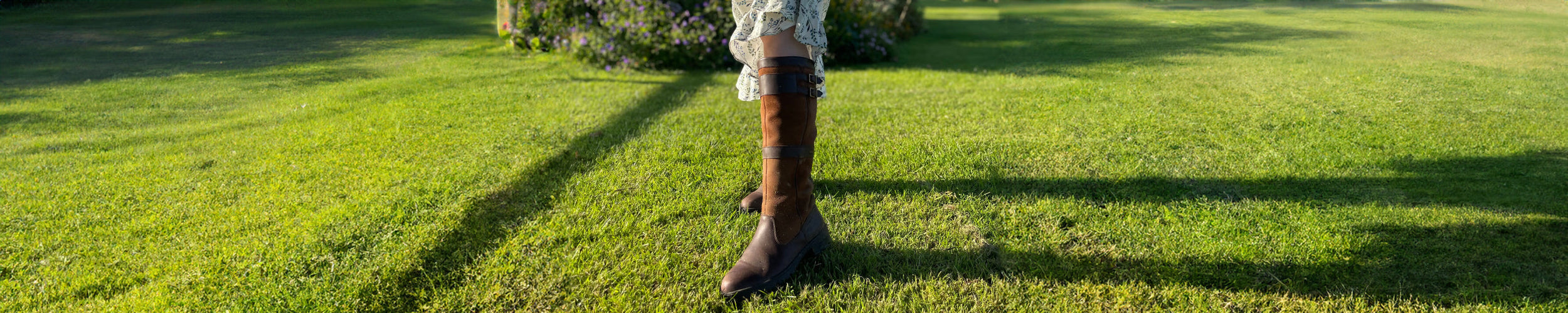 Wide image of the Dubarry Longford Leather Waterproof and Breathable Country Boot being worn with a dress on a well-maintained lawn with some shrubs in the background.